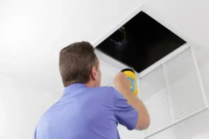 A person shines a flashlight into a dark ceiling vent, meticulously inspecting it for any signs of needed air duct repair.