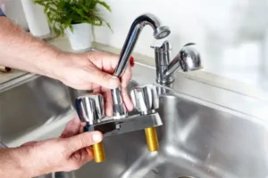 Hands expertly grasp a shiny, detached kitchen faucet above a stainless steel sink. In the background, another faucet stands ready for its role in kitchen plumbing installation, accompanied by a verdant plant adding a touch of nature.