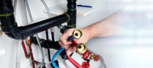 Person holding red and blue hoses in front of plumbing pipes in a utility space.