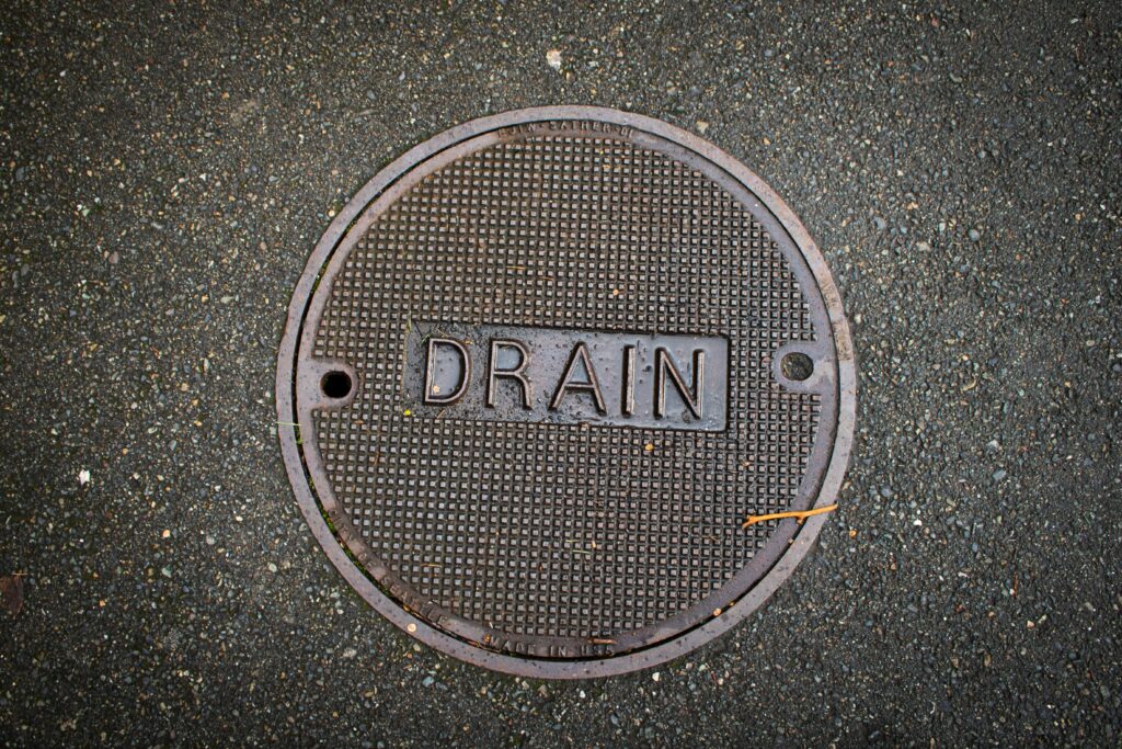A round metal manhole cover labeled "DRAIN" set in asphalt pavement.